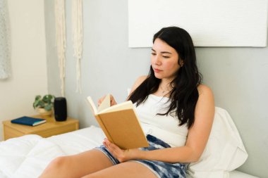 Woman in her 30s enjoys reading a book while relaxing comfortably on her bed in a brightly lit, cozy bedroom