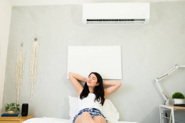 Beautiful woman enjoying the cool comfort of a mini split air conditioning unit on a hot summer day while relaxing in a bed