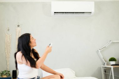 Caucasian woman adjusting a mini split air conditioning unit with a remote control while sitting comfortably on her bed on a hot summer day