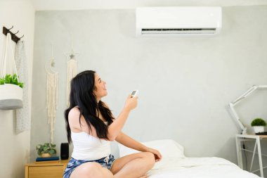Young woman relaxes on her bed with a remote in hand, enjoying the cool summer atmosphere provided by the mini split air conditioning unit