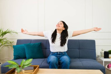 Beautiful woman lounges on a sofa in a sunlit, inviting living room in her new home, stretching her arms wide and radiating satisfaction