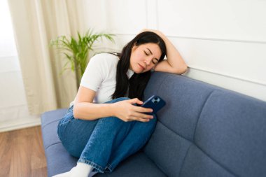 Young woman sitting on her sofa looking at her mobile phone and expecting a call or a message at home