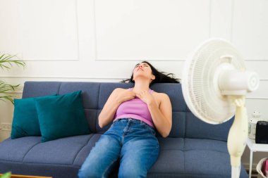 Woman trying to cool off in her home during the hot summer with the help of a fan, expressing discomfort from heat
