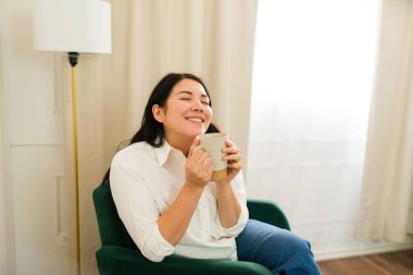 Joyful woman savoring a warm cup of coffee while relaxing in her bright, cozy living room, exuding contentment and comfort