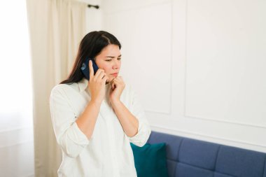 Young woman appears troubled while talking on her phone, standing anxiously in a minimalist, well-lit living room