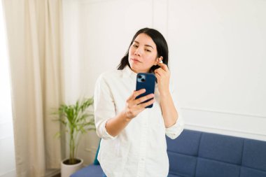 Young woman in using her smartphone  with earbuds and getting ready to take a phone call in a brightly lit home living room