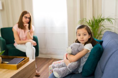Cute little girl making eye contact while hugging a teddy bear during a therapy session with a child psychologist