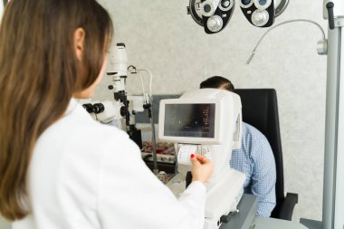 Bir klinikteki oftalmolog görme değerlendirmesi için özel teşhis ekipmanları kullanan bir erkek hastaya detaylı bir göz kontrolü yapar.