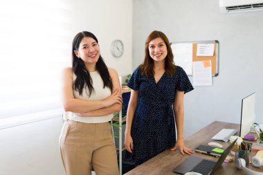 Parlak bir ofis ortamında güvenle duran iki neşeli bayan girişimci iş ortaklığı ve takım çalışması tasvir ediyorlar.