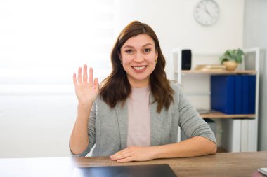 Parlak bir ofis ortamında masasında el sallayarak gülümseyen profesyonel bir kadın.