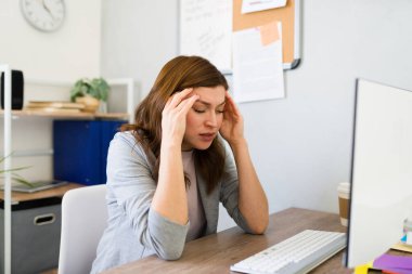 Genç iş kadını, modern bir ofis ortamında masasında çalışırken bunalmış ve stresli hissediyor.