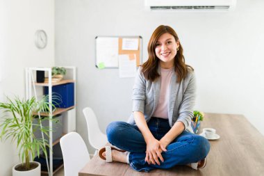 Modern bir çalışma ortamında ofis masasında bağdaş kurmuş, güler yüzlü bir kadın güven ve rahatlık içinde oturuyor.