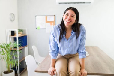 Profesyonel ve yaklaşılabilir iş ortamı. Modern bir ofiste oturan mavi gömlekli, gülümseyen genç bir kadın tarafından temsil ediliyor.