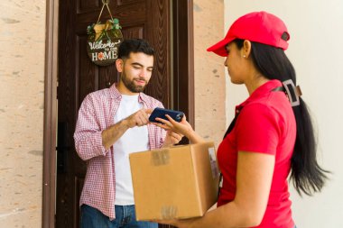 Müşteri evin girişinde dijital imza için akıllı telefon kullanırken İspanyol teslimatçı kadın elinde bir paket tutuyor.