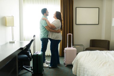 Hoş bir çift, bir yolculuktan sonra bir otel odasında, bavullarla çevrili, sevgi ve mutluluk saçan bir şekilde kucaklaşır. Hafta sonu kaçamakları için günlük giysiler giyip romantik bir anı paylaşıyorlar.