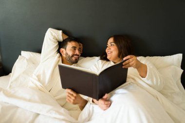 Mutlu çift otelde kalmaktan zevk alıyor ve yatakta yatarken oda servisi menüsünü kontrol ediyor.