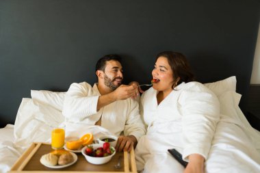 Beyaz bir adam otel odasında yatakta kahvaltı ederken kız arkadaşına çilek yediriyor.