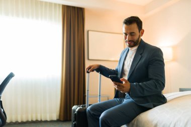 Bir otel odasına vardıktan sonra elinde bavulla yatakta oturan ve akıllı telefon kullanan mutlu bir işadamı.