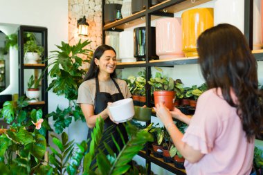 Neşeli satıcı kadın, hoş bir bitki dükkanında müşteriye saksıları sunar, bitki meraklıları için ideal rahat bir ortam yaratır.