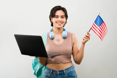 Okulda İngilizce öğrenirken elinde dizüstü bilgisayarla Amerikan bayrağını sallayan neşeli genç bir kızın portresi.