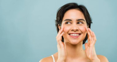 Mükemmel bir gülüşü olan genç bir kadın kameraya bakmadan yüzüne dokunuyor, mutluluğu ve mutluluğu ifade ediyor.