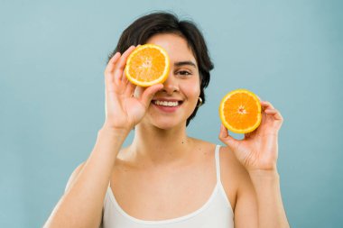 Gülümseyen kadın bir stüdyo çekiminde gözlerinin üzerinde dilimlenmiş portakallar tutuyor, mutluluk saçıyor ve sağlıklı cilt bakımını sergiliyor.