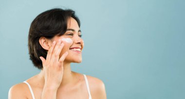 Şirin bir kadın yüzüne nemlendirici krem sürüyor ve stüdyo çekimlerinde gülümsüyor.