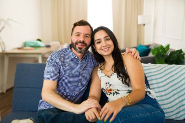 Oturma odasındaki rahat bir kanepede rahatlarken el ele tutuşup gülümseyen çok ırklı genç bir çift.