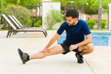 Güneşli bir günde havuz başında egzersiz yaptıktan sonra spor kıyafetleriyle bacakları esneten odaklanmış erkek sporcu.