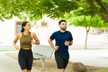Genç İspanyol çift birlikte bir şehir parkında koşuyor, gülümsüyor ve açık havada güneşli bir günün tadını çıkarıyor, aktif yaşam tarzlarını ve spor yapmaya bağlılıklarını sergiliyorlar.