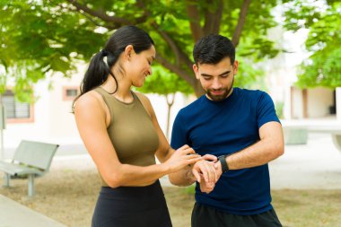 Gençliklerinde atletik kıyafetler giyen İspanyol bir çift parkta spor salonunda spor yaparken spor takip cihazını kontrol ediyor.