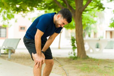 Sporcu acı içinde kıvranıyor, sakat dizini sakin bir parkta egzersiz yaparken tutuyor. Rahatsızlığa rağmen, etkin bir yaşam tarzının zorluklarını vurgulayarak zorluklara göğüs geriyor.