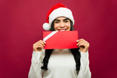 Gülümserken ve Noel Baba şapkası takarken ağzında boş kırmızı bir hediye kartı tutan neşeli bir kadın.