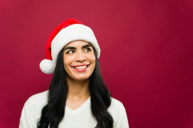 Güzel bir kadın Noel Baba şapkası takıyor ve büyük bir gülümsemeyle soluna bakıyor.