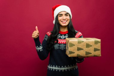 Noel Baba şapkası takan ve kışlık süveter giyen Latin bir kadın elinde Noel hediyesi tutuyor ve kırmızı arka planda baş parmak hareketi yapıyor.