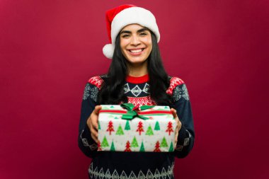 Muhteşem bir kadın elinde Noel Baba şapkası ve rahat bir süveter giyiyor, kırmızı bir fona gülümsüyor.