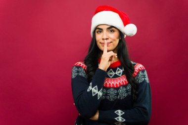 Noel Baba şapkası takan güzel Latin bir kadın Noel arkaplanındaki parmaklarıyla sessiz bir jest yapıyor.
