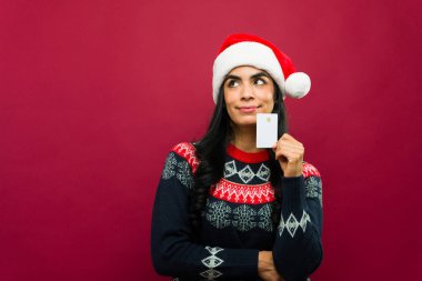 Noel Baba şapkası takan düşünceli bir kadın elinde bir kredi kartı tutuyor ve Noel alışveriş listesini düşünüyor.