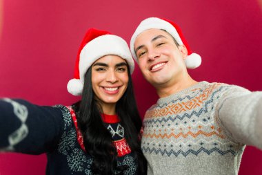 Noel Baba şapkası ve çirkin Noel süveteri giyen genç Latin çift kırmızı arka planda selfie çekiyorlar.