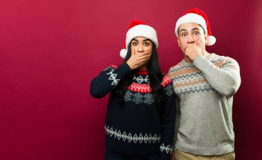 Noel süveteri ve Noel Baba şapkası giyen genç bir çift ağızlarını elleriyle kapatıyor ve kırmızı arka planda şaşırmış görünüyorlar.