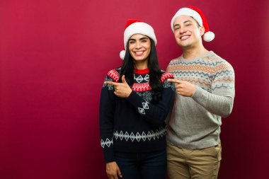 Gençliklerindeki İspanyol çift Noel Baba şapkaları ve Noel süveterleri giyerek kırmızı arka plandaki fotokopi alanını gösteriyorlar.