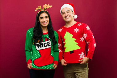 Gülünç ve çirkin Noel süveterleri içinde neşeli kırmızı bir arka planda gülümseyerek poz veren mutlu genç çift.