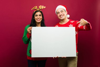 Mutlu ve neşeli bir çift Noel süveteri giyip kırmızı arka planda boş bir tabela tutuyorlar.