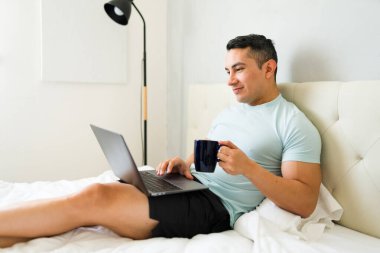 Yirmili yaşlardaki yakışıklı bir adam bir fincan kahvenin tadını çıkarıyor ve yatağının rahatlığından laptopunu kullanıyor.