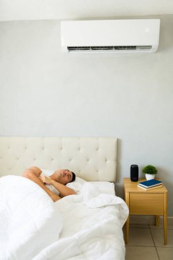 Rahat bir adam sıcak bir yaz günü yatağında rahat bir şekilde uyurken klimasının serin havasının tadını çıkarıyor.