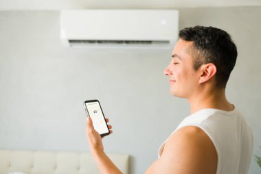 Rahatlamış latin adam yatak odasında dururken akıllı telefon uygulaması ile mükemmel oda sıcaklığını ayarlıyor.