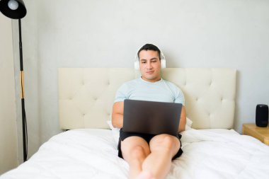 Genç adam evde çalışıyor, laptopuyla yatağında oturuyor ve kulaklık takıyor.