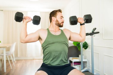 Kas yığını olan bir adam, kişisel fitness sahasında bir bankta otururken, pazılarını halterlerle çalıştırıyor.