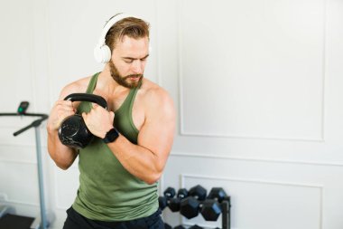 Formda bir adam spor yaparken ve spor salonunda müzik dinlerken bir kettlebell kaldırıyor.