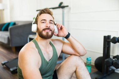 Atletik genç adam evde egzersiz yaptıktan sonra bir mola verip kulaklıkla müzik dinliyor.